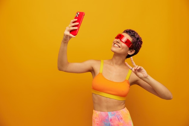 Femme blogueuse souriante dans des lunettes millénaires inhabituelles prenant des selfies sur son téléphone en tenue de sport sur un fond de studio orange espace libre