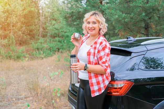 Une femme blonde adulte boit du thé dans un thermos dans la nature debout à côté d'une voiture