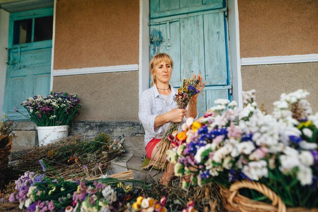 Une femme blonde adulte est assise dans le pays et crée des bouquets de fleurs séchées cultivées dans le jardin