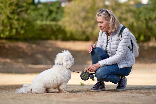 Femme blonde adulte marchant avec un chien blanc moelleux dans le parc de la ville.