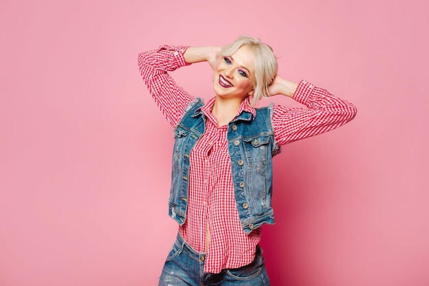 Femme blonde adulte très positive et heureuse vêtue d'une chemise et d'un jean bleu posant sur fond rose