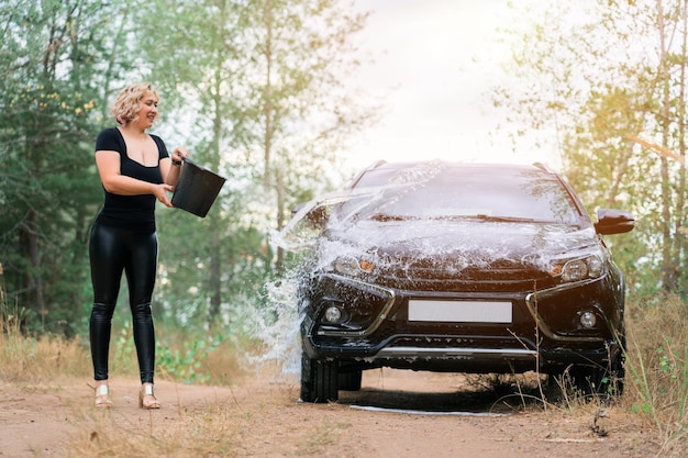Une femme blonde d'âge moyen arrose une voiture avec un seau d'eau