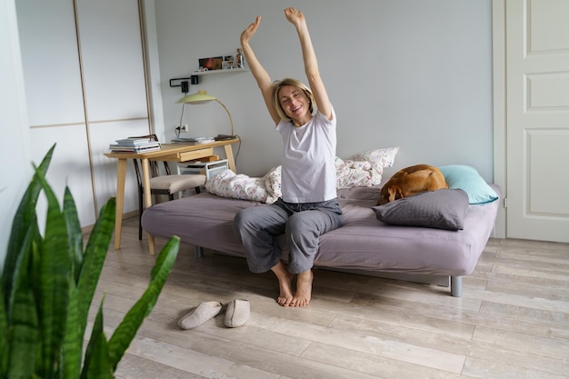 Une femme blonde d'âge moyen a commencé sa matinée au lit avec un chien et s'étire avec un sourire de plaisir