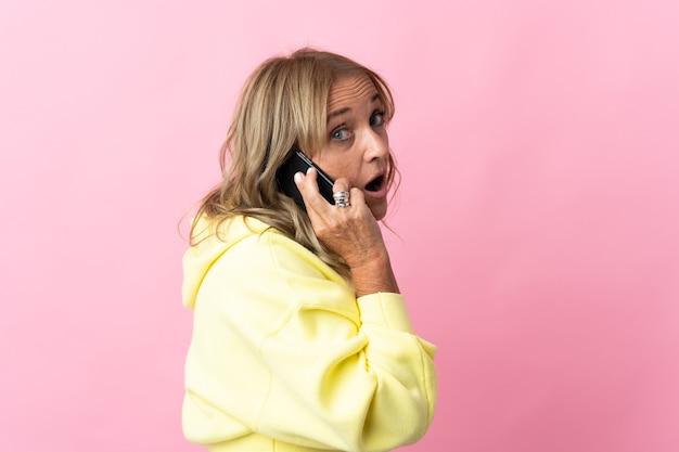 Femme blonde d'âge moyen sur mur rose isolé en gardant une conversation avec le téléphone mobile avec quelqu'un