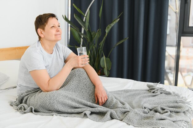 Femme blonde d'âge moyen souriante heureuse buvant un verre d'eau allongé sur le lit à la maison