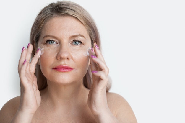 Une femme blonde âgée se masse le visage avec une crème pendant les procédures de spa sur un mur blanc