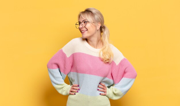 Femme blonde à l'air heureuse, joyeuse et confiante, souriante fièrement et regardant de côté avec les deux mains sur les hanches