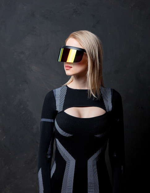 Femme blonde androïde dans des lunettes VR et costume sur fond sombre.