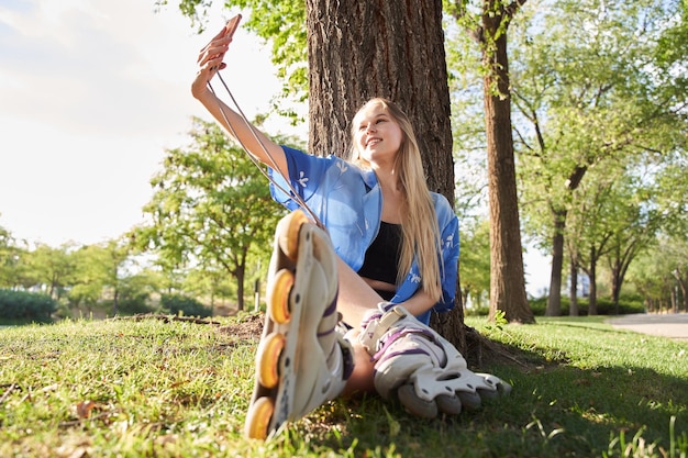 Femme blonde assise sur un arbre avec des patins à roues alignées en prenant un selfie avec son téléphone intelligent