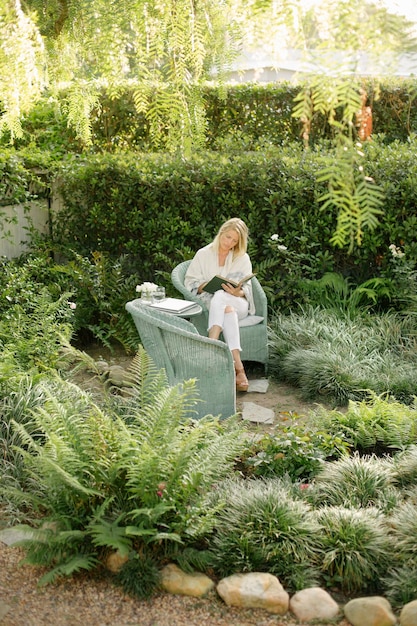 Femme blonde assise sur une chaise en osier dans un jardin lisant