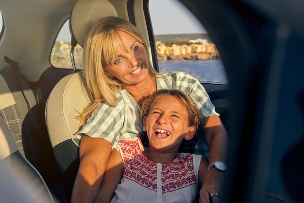 Femme blonde assise dans une voiture avec sa fille allongée sur elle tout en riant avec la mer en arrière-plan