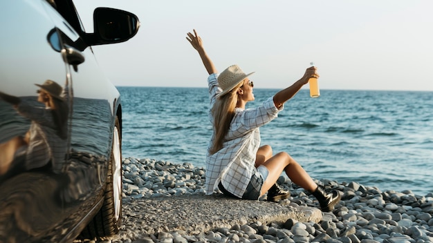 Femme blonde assise sur des rochers avec du jus en regardant la mer près de la voiture