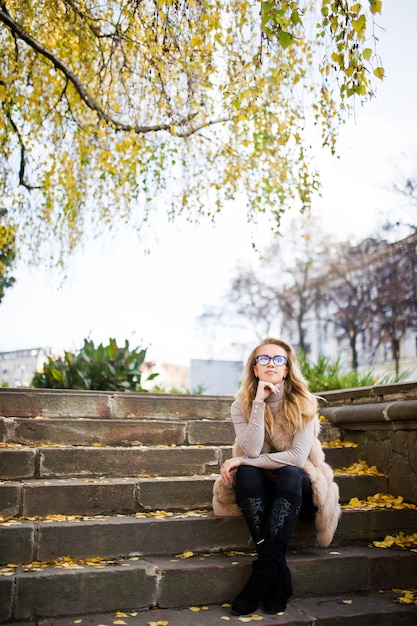 Femme blonde au manteau de fourrure et lunettes, assis dans les escaliers avec des feuilles jaunes.