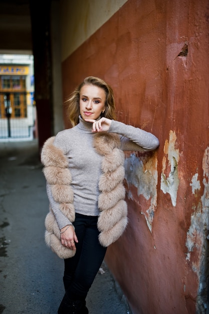 Femme blonde au manteau de fourrure posée contre le vieux mur orange.