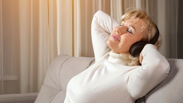 Femme blonde aux cheveux longs d'âge moyen avec une frange portant un col roulé léger écoute de la musique via des écouteurs sans fil se relaxant sur la lumière du soleil du canapé