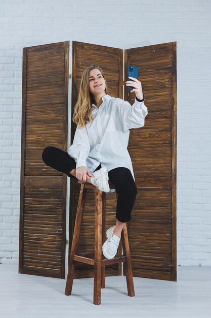 Femme blonde aux cheveux longs dans une chemise blanche et un pantalon noir posant devant la caméra du téléphone tenant le téléphone et prenant un selfie sur un fond isolé
