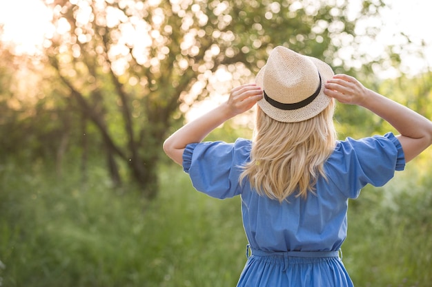 Femme blonde en belle robe bleue tenant un chapeau de paille dans ses mains et s'amusant dans le parc