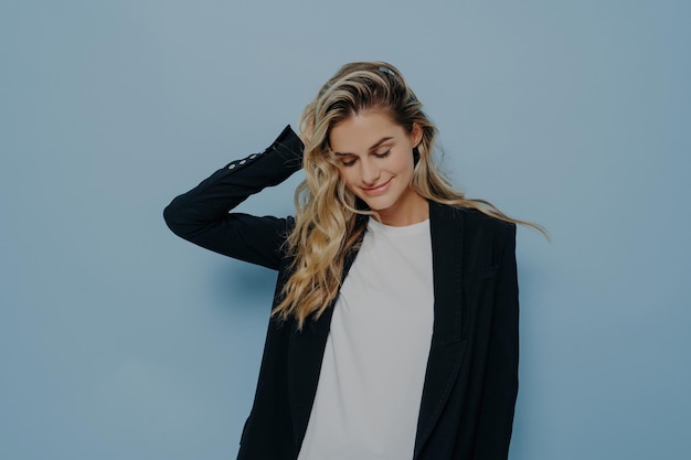 Femme blonde en blazer noir et t-shirt blanc sourit avec les yeux fermés isolés sur fond bleu Vibes positives