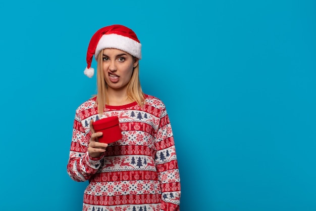 Femme blonde avec bonnet de Noel se sentant dégoûté et irrité, tirant la langue, n'aimant pas quelque chose de méchant et dégueulasse