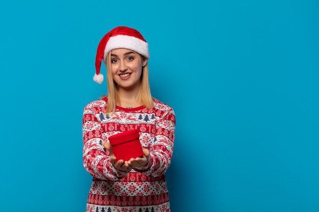 Femme blonde avec bonnet de noel souriant joyeusement avec un regard amical, confiant et positif, offrant et montrant un objet ou un concept