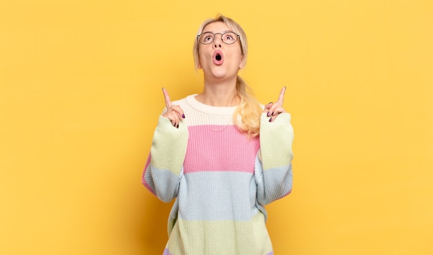 Femme blonde à la bouche choquée, étonnée et ouverte, pointant vers le haut avec les deux mains pour copier l'espace