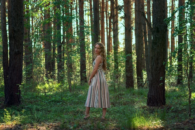 Femme blonde bouclée dans une robe se promène dans une forêt de pins