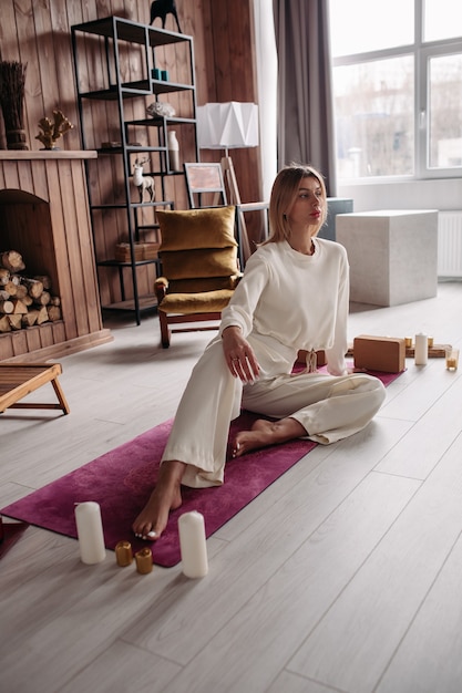 Une femme blonde caucasienne en blanc se reposant sur un tapis rose vif, pieds nus, entourée de bougies prêtes pour la méditation à la maison.