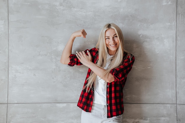 Une femme blonde caucasienne excitée en chemise à carreaux montre des biceps contre un mur de marbre satisfaite