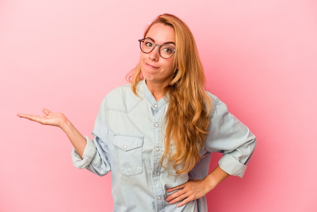 Femme blonde caucasienne isolée sur fond rose montrant un espace de copie sur une paume et tenant une autre main sur la taille.