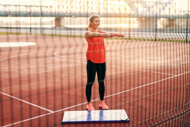 Femme blonde caucasienne souriante, faire des exercices de fitness avec des haltères en se tenant debout à l'extérieur.