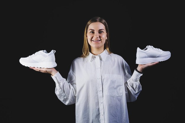 Une femme blonde en chemise blanche sur fond noir tient dans ses mains des baskets blanches en cuir véritable