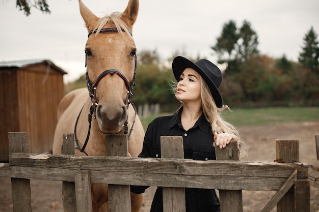 Femme blonde et cheval brun debout dans une ferme. Femme portant des vêtements noirs et un chapeau. Femme touchant le cheval derrière la clôture.