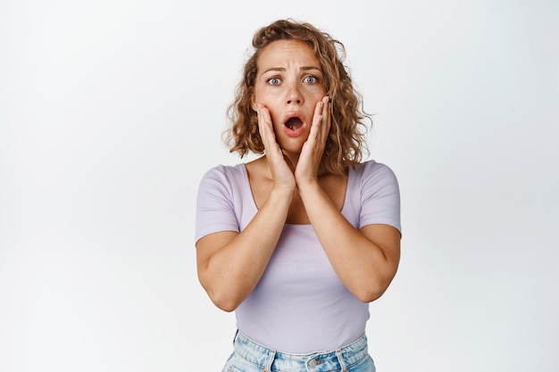 Une femme blonde choquée et inquiète halete, l'air surprise devant la caméra, témoin d'une catastrophe, debout sur fond blanc.