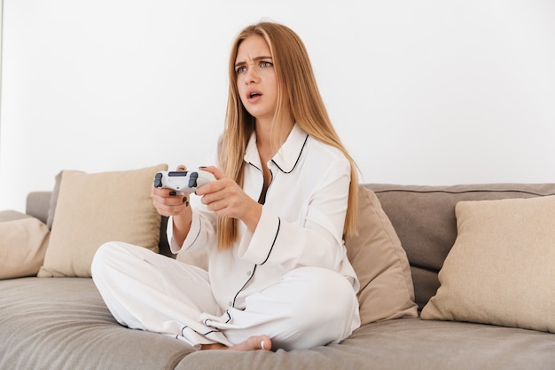 Femme blonde confuse en pyjama jouant à un jeu vidéo assis sur un canapé dans un salon lumineux