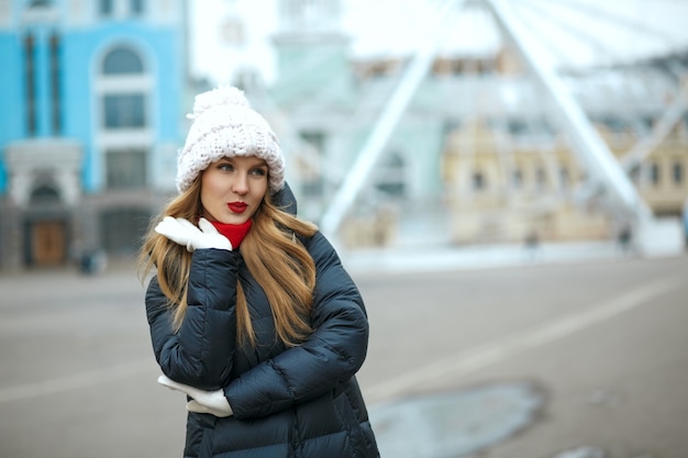 Femme blonde coquette avec du rouge à lèvres portant une casquette tricotée, profitant d'une promenade en ville. Espace pour le texte