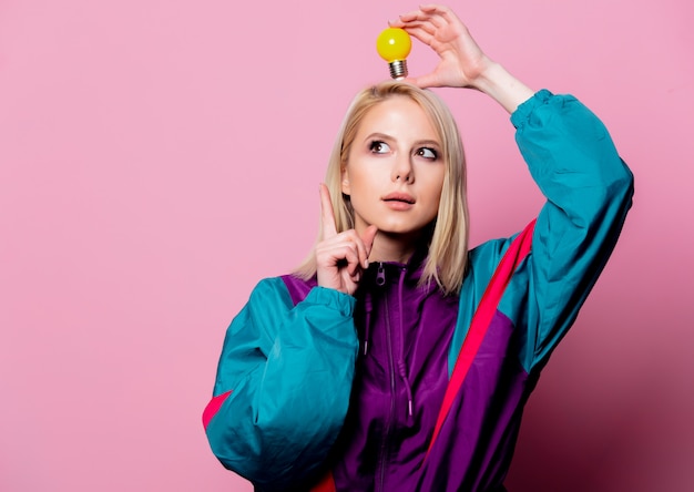 Femme blonde en costume de sport des années 90 avec ampoule sur mur rose