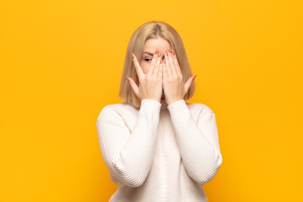 Femme blonde couvrant le visage avec les mains, furtivement entre les doigts avec une expression surprise et regardant sur le côté