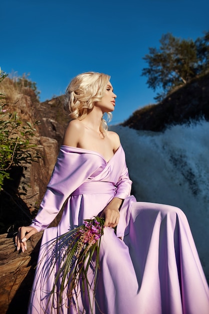 Femme blonde dans une longue robe rose assise sur une pierre près de la cascade. Magnifiques cheveux longs beau sourire sur le visage de la fille
