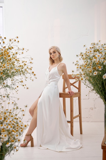 Une femme blonde dans une robe de mariée blanche debout à la maison parmi les bouquets de fleurs sauvages