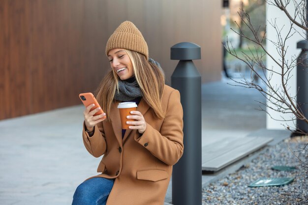 Femme blonde discutant sur Internet en ligne sur smartphone pendant la pause-café tout en se relaxant sur un banc de parc