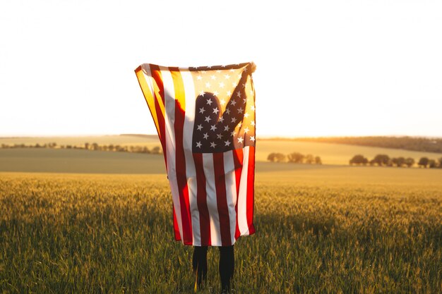 Femme blonde avec le drapeau américain en cours d'exécution dans un champ de blé au coucher du soleil. Fête de l'indépendance, fête patriotique. 4 juillet.
