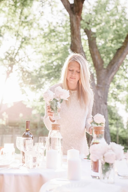 Femme blonde dressant une table dans un jardin des bougies et des vases avec des roses roses