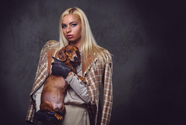 Une femme blonde élégante vêtue d'une veste à l'ancienne tient un chien blaireau rouge.