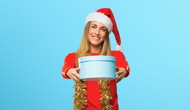 Femme blonde habillée pour les vacances de Noël tenant des coffrets cadeaux dans les mains sur fond bleu