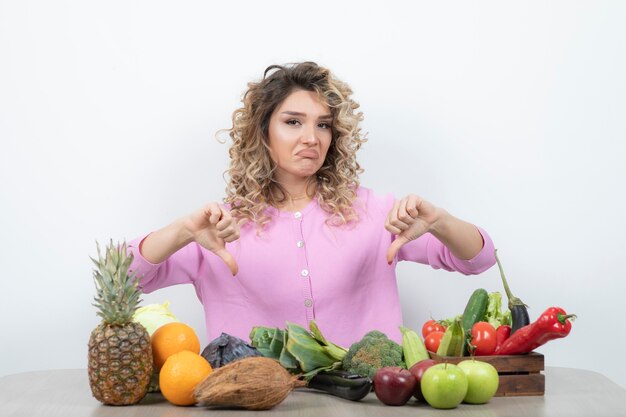 Femme blonde en haut rose assis à table avec de nombreux fruits donnant les pouces vers le bas.