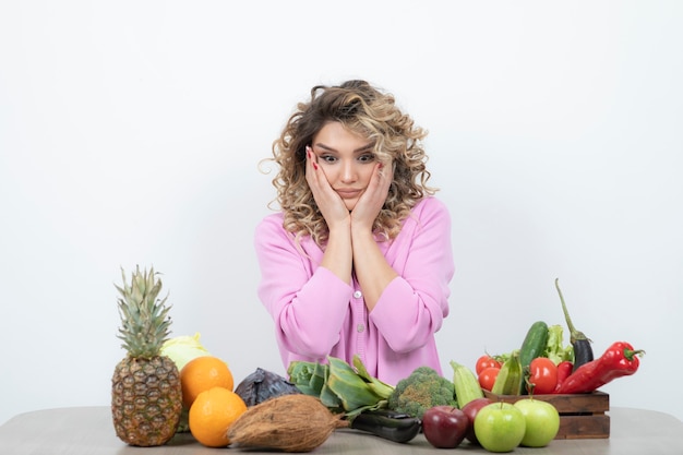 femme blonde en haut rose assis à table avec de nombreux fruits et légumes.