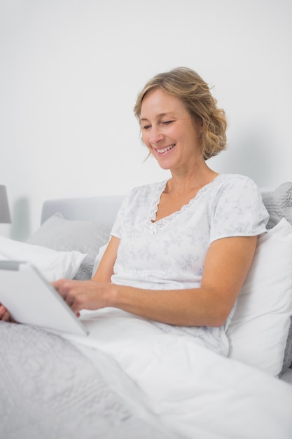 Femme blonde heureuse assis dans son lit à l&#39;aide de tablet pc