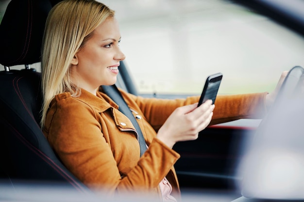 Une femme blonde heureuse est assise dans sa voiture et utilise le téléphone
