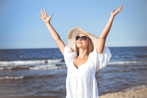 Une femme blonde heureuse est sur la plage de l'océan dans une robe blanche, des lunettes de soleil et un chapeau, levant les mains.