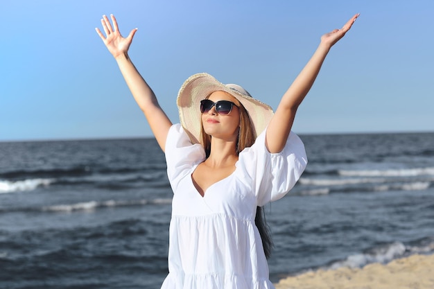 Une femme blonde heureuse est sur la plage de l'océan dans une robe blanche, des lunettes de soleil et un chapeau levant les mains